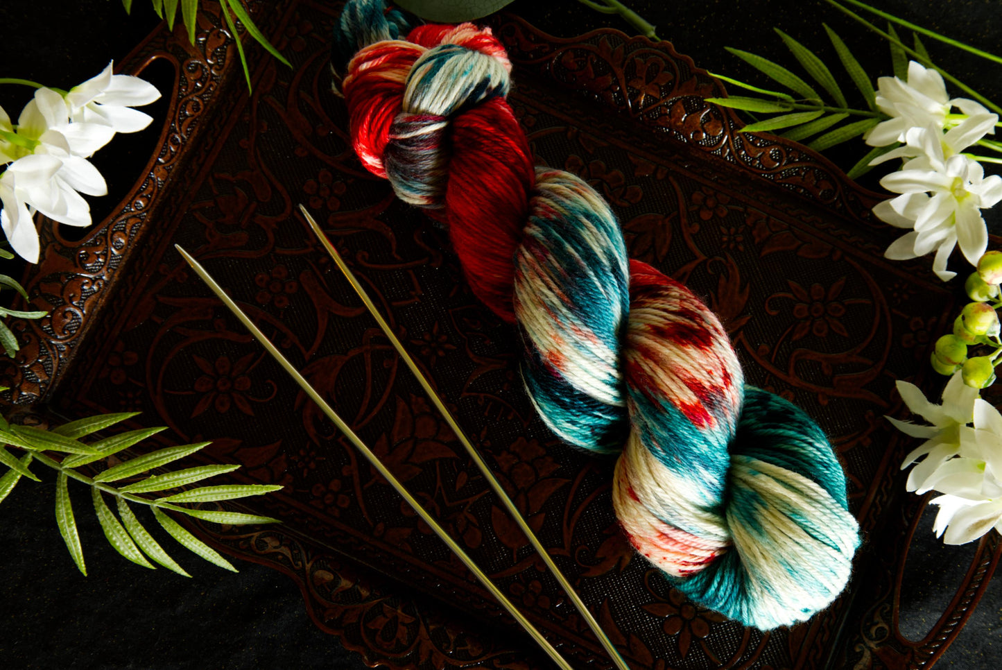 A skein of yarn lays on a copper tray next to a set of silver knitting needles, surrounded by flowers. The yarn is hand dyed red, green, and white.