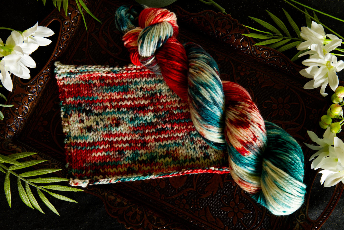 A skein of hand dyed yarn lays next to a swatch that details the color placement once the yarn is knit. It is hand dyed red, green, and white. They are surrounded by flowers.