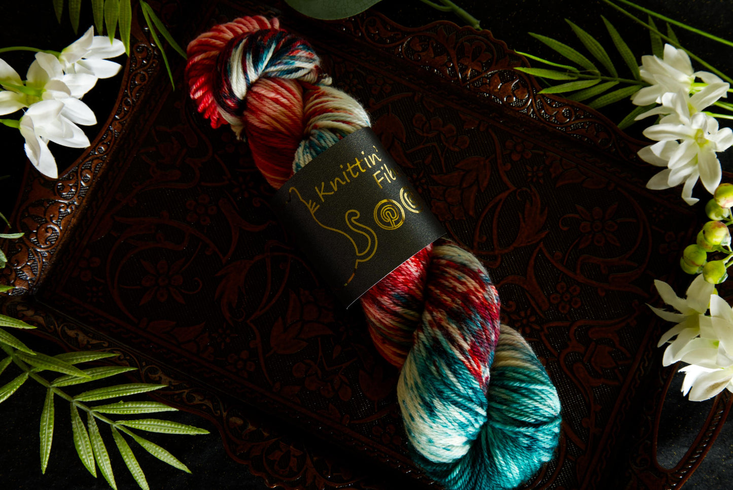 A single skein on yarn lays on a copper tray surrounded by flowers. It is hand dyed red, green, and white. The yarn is twisted into a skein and wrapped in a product label that says "Knittin' Kitten Fiber."