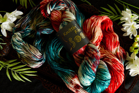 Skein of hand dyed yarn that is red, white, and green. It is wrapped in a product label by Knittin' Kitten Fiber. The yarn lays on a table and is surrounded by flowers. 