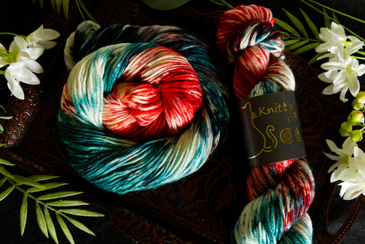 Two skeins of hand dyed yarn are shown. One is wound into a circle and the oher is twisted into a skein with a product label reading "Knittin' Kitten Fiber." The yarn is red, green, and white. It lays on a table surrounded by flowers. 
