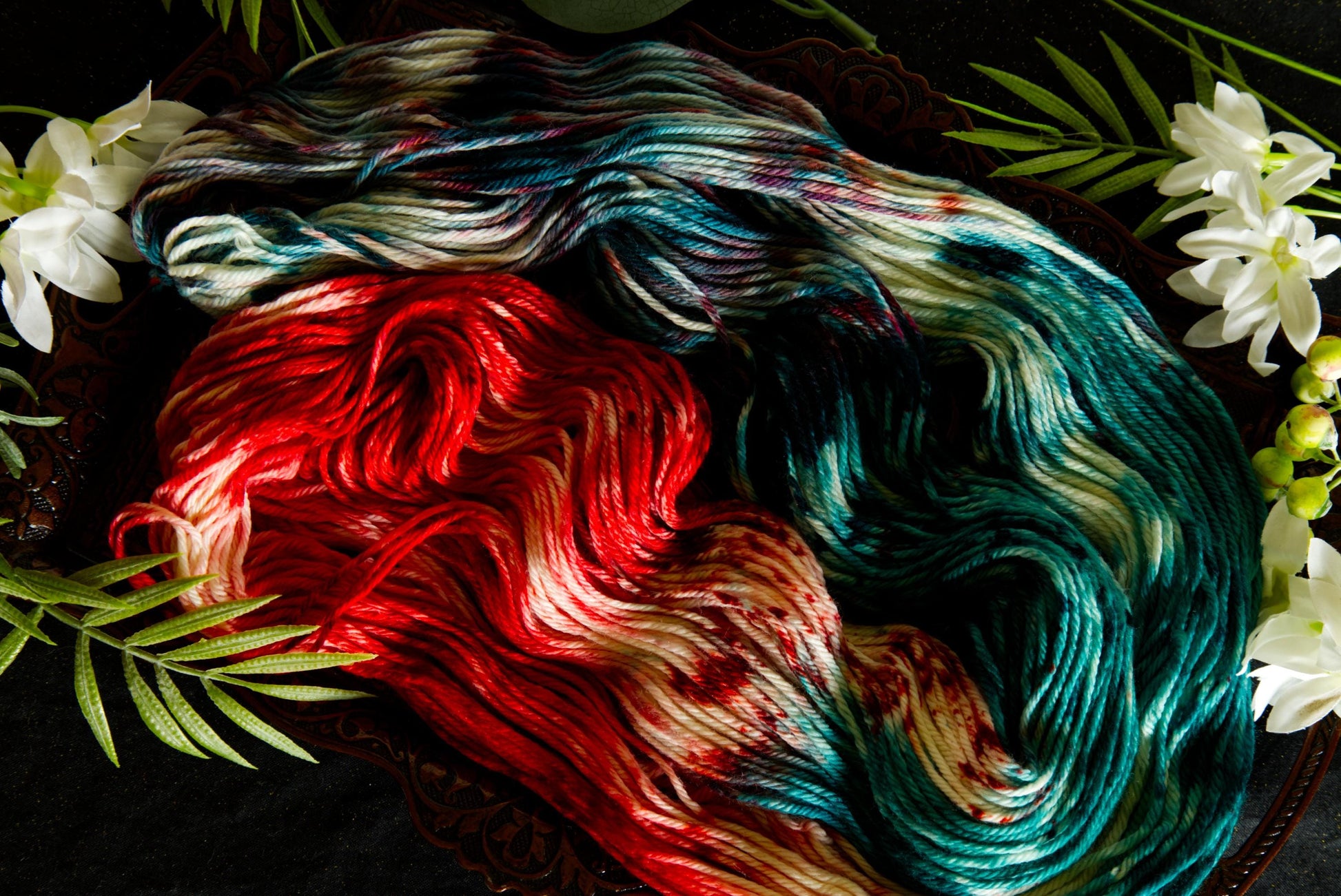 A hank of hand dyed yarn lays spread out on a table, surrounded by flowers. It is red, green, and white. 